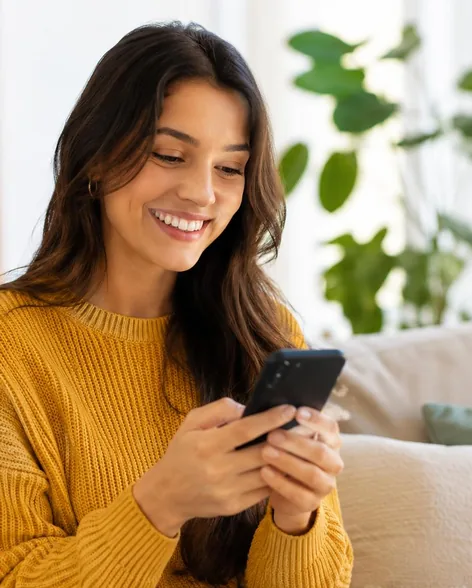 Mujer sonriendo mientras usa su celular en el living de su casa